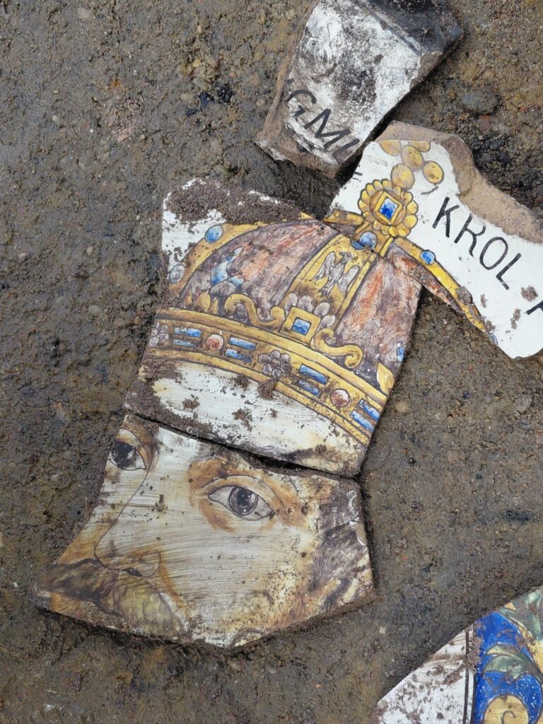 Fragmenty ceramiczne z malowidłem przedstawiającym twarz króla, częściowo pokryte ziemią. Widoczne napisy „KRÓL” i „CM”.