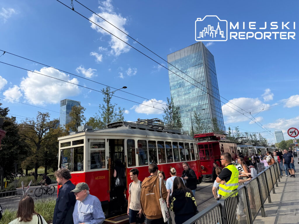 Tłum ludzi obserwuje tramwaje podczas Mistrzostw motorniczych na ulicy w otoczeniu nowoczesnych biurowców.