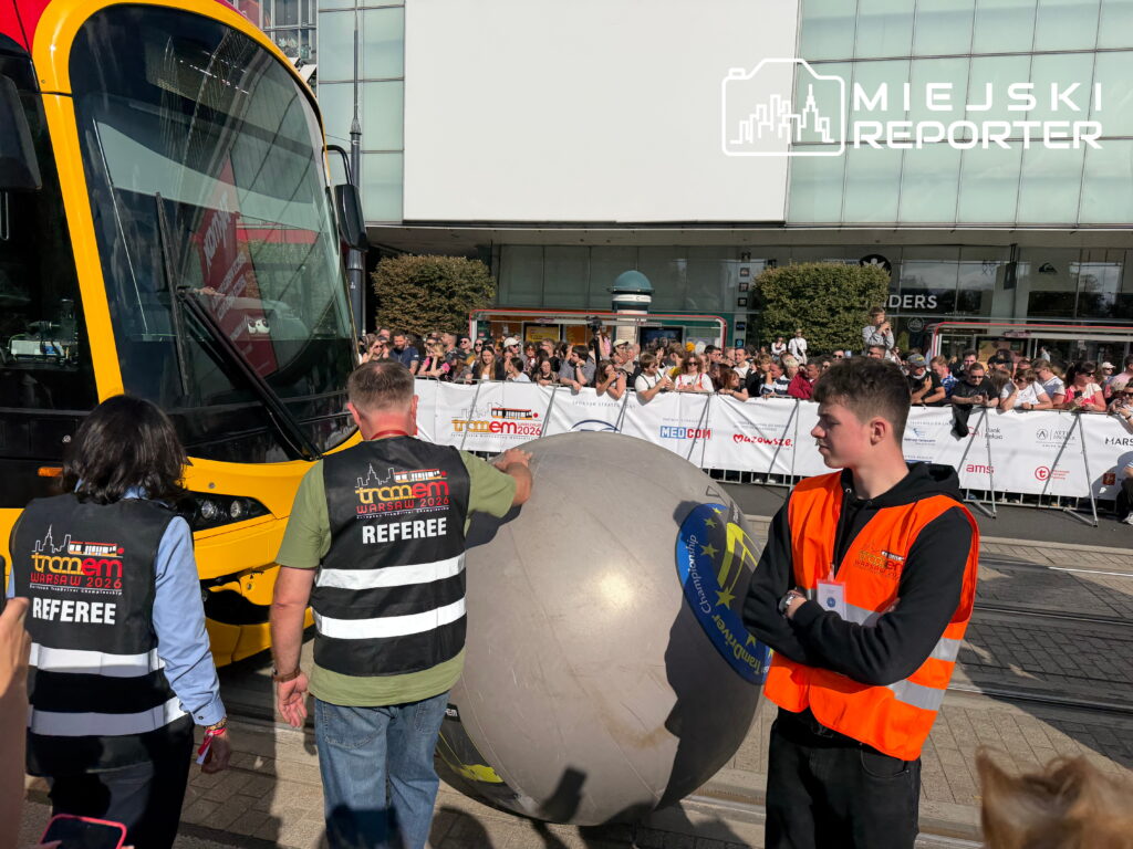 Mężczyźni w kamizelkach sędziów przygotowują dużą kulę do zawodów, a w tle widoczna jest tłumnie zgromadzona publiczność podczas Mistrzostw motorniczych.