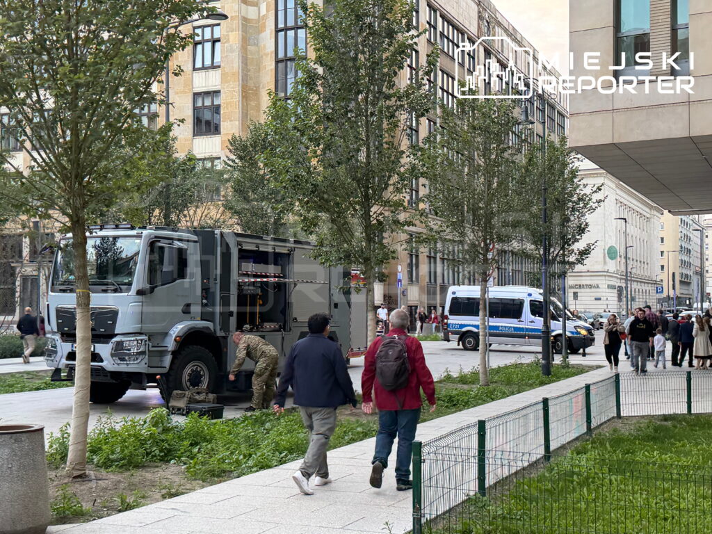 Żołnierz przygotowuje sprzęt obok opancerzonego pojazdu w miejskiej przestrzeni z przechodniami i drzewami w tle.