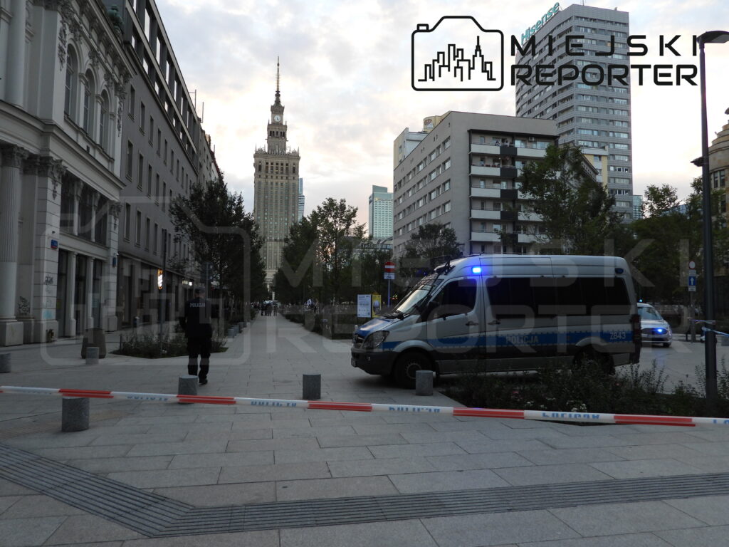 Policjant stojący przy policyjnym vanie z włączonymi sygnałami świetlnymi na ulicy, z widokiem na wysokie budynki w tle.