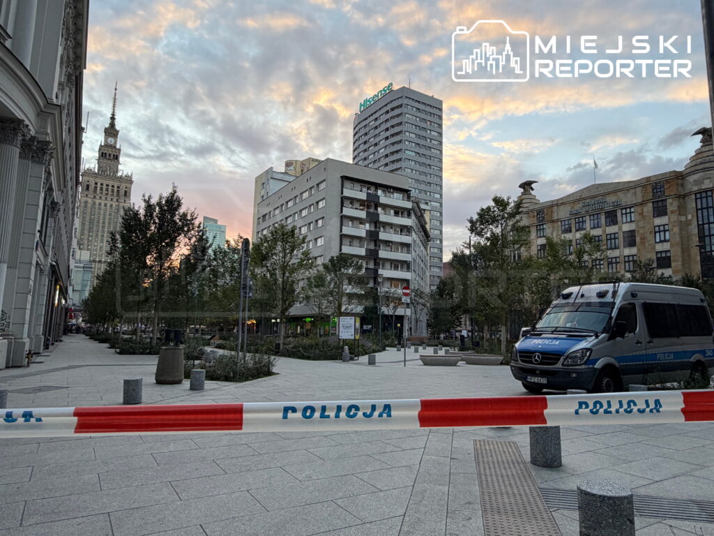 Policyjny radiowóz stoi na placu, otoczony taśmą z napisem "POLICJA", w tle widoczny nowoczesny biurowiec.