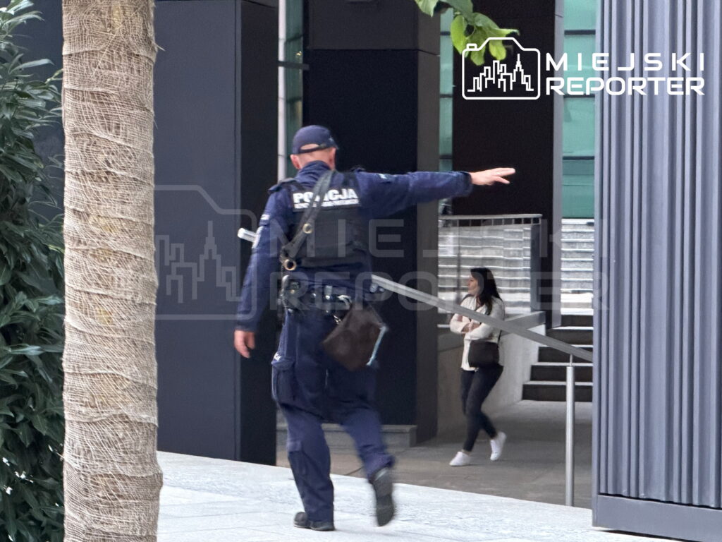 Policjant w mundurze wskazuje kierunek, przechodząc obok nowoczesnego budynku, obok którego stoi kobieta.