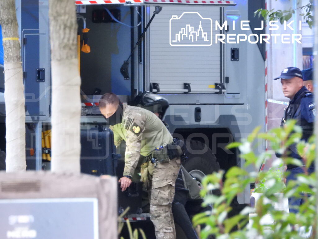 Funkcjonariusz w mundurze wojskowym przeszukuje sprzęt w pojeździe służbowym, obok stoi policjant.