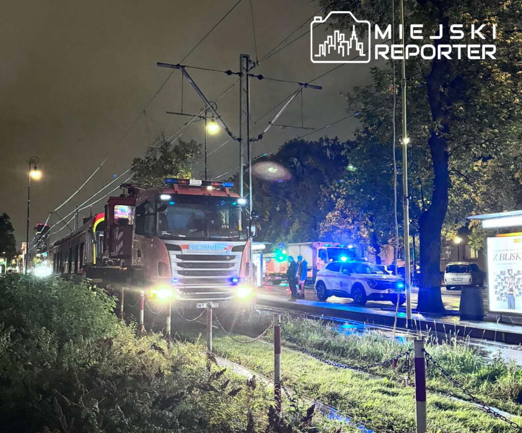Czerwony pojazd ciężarowy z logo na boku stoi obok torów tramwajowych, a w tle widać policyjne radiowozy z włączonymi sygnałami.