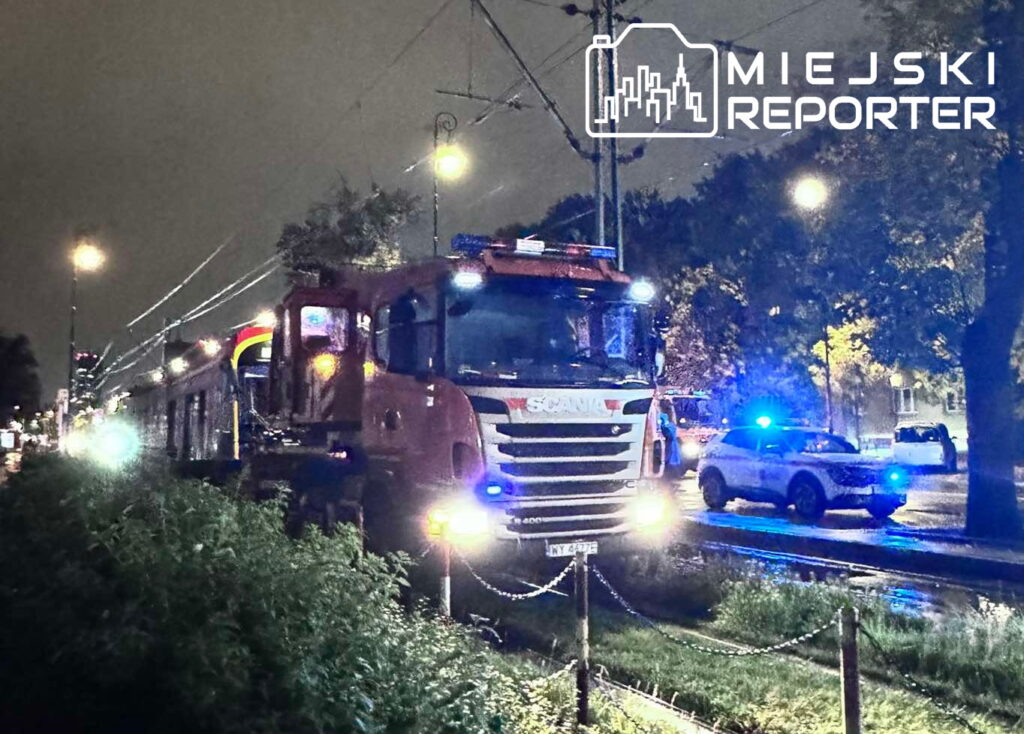 Czerwony wóz strażacki z oświetleniem awaryjnym stoi obok torów tramwajowych w deszczowy wieczór.