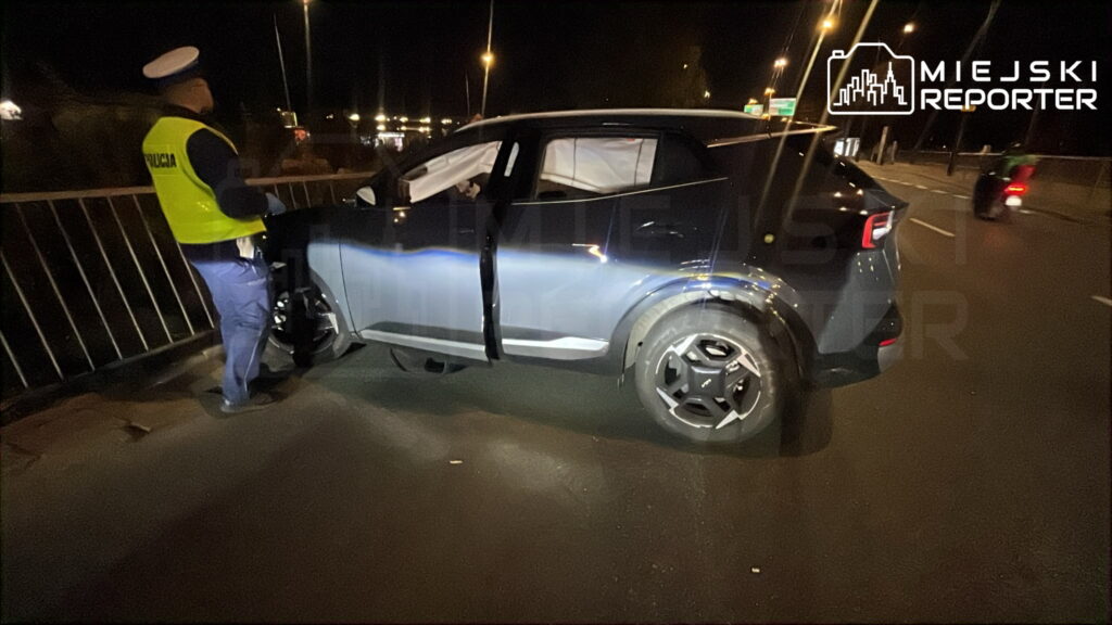 Policjant w odblaskowej kamizelce sprawdza uszkodzoną Kia, która uderzyła w barierki na drodze nocą.