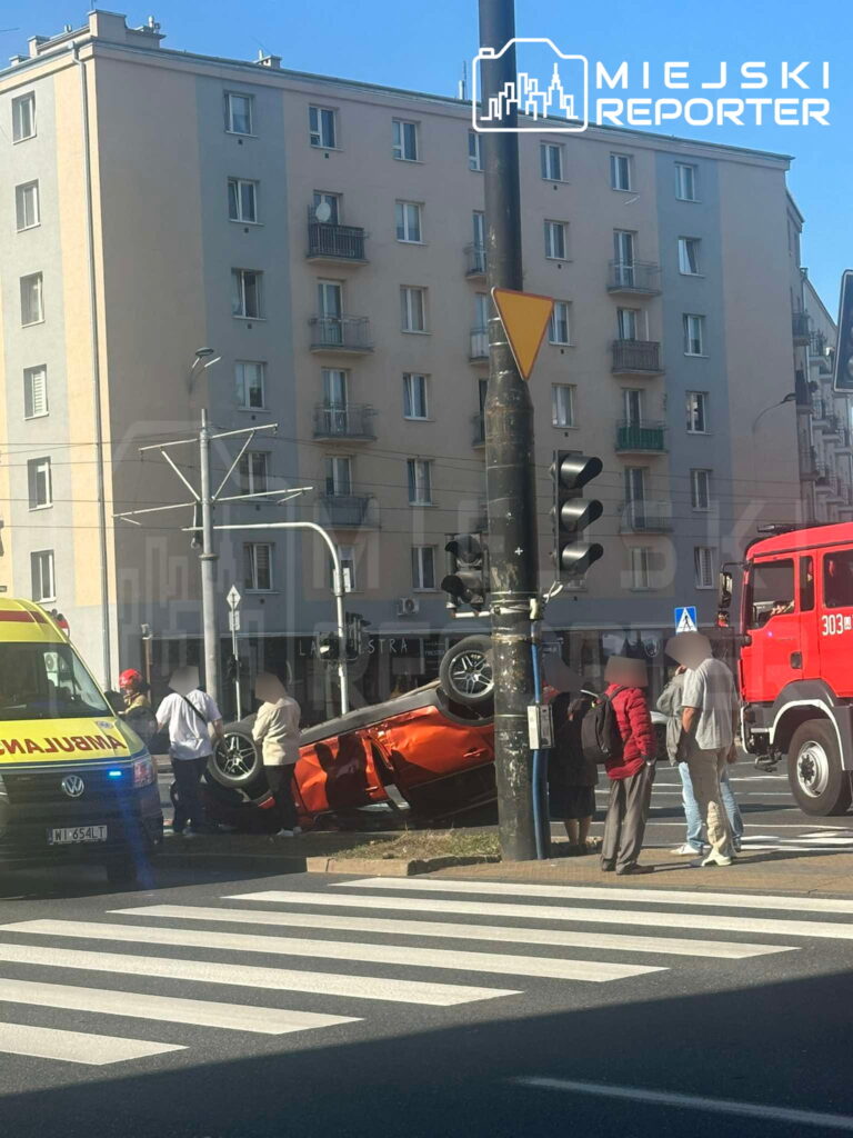 Czerwony samochód osobowy dachował obok sygnalizacji świetlnej, a obok znajdują się strażacy i ratownicy medyczni.