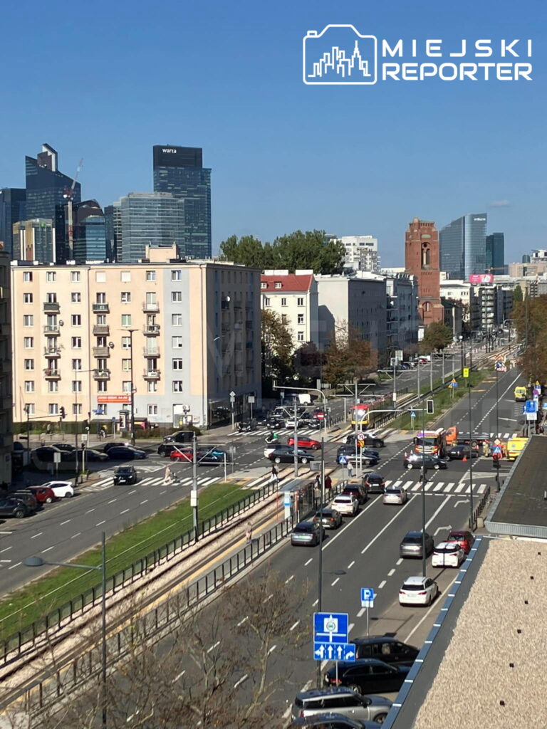 Ruch samochodowy na ulicy z widokiem na nowoczesne biurowce i zabytkową wieżę, w tle niebieskie niebo.
