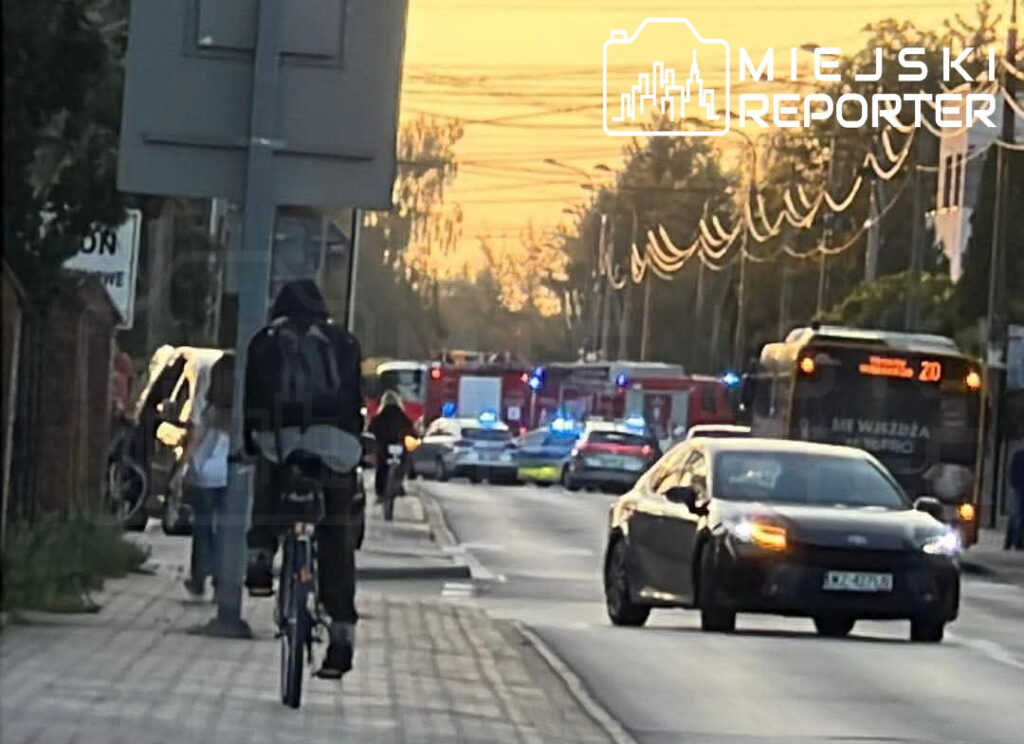 Rowerzysta przejeżdża obok miejsca wypadku na Arkuszowej, gdzie interweniują służby ratunkowe z włączonymi sygnałami.