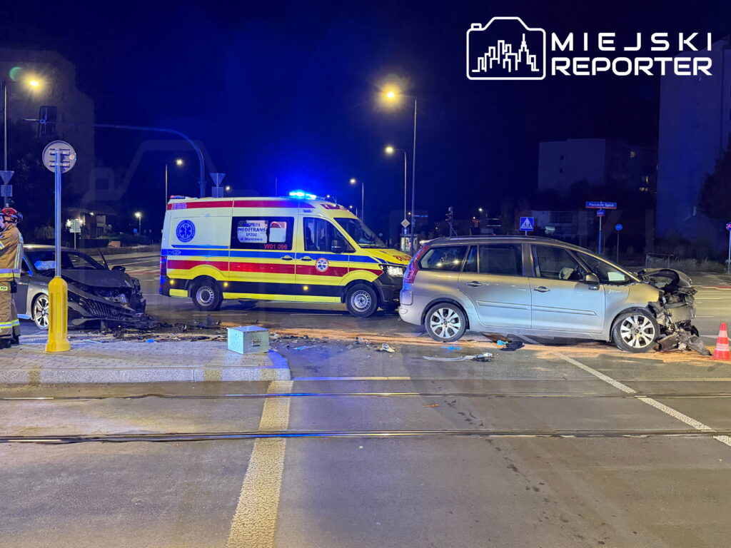 Uszkodzony szary minivan i czarny samochód po zderzeniu na skrzyżowaniu, obok karetka i strażak w nocy.