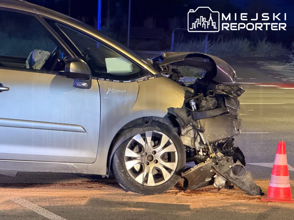 Srebrny Citroen z rozbitym przodem stoi na jezdni nocą, obok uszkodzonego auta i pachołka drogowego.