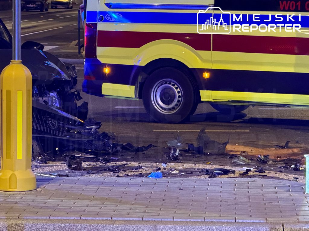 Rozbity samochód i porozrzucane szczątki na jezdni obok karetki pogotowia po zderzeniu drogowym