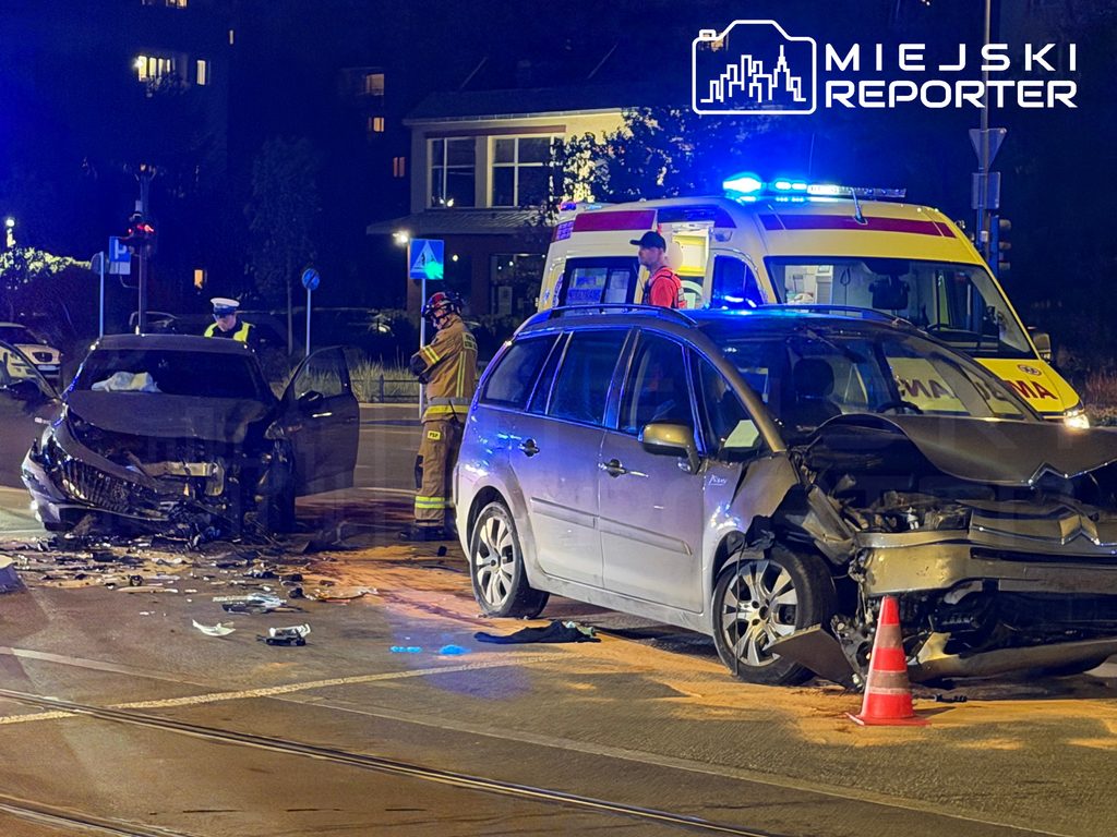 Dwa rozbite samochody na ulicy nocą, obok karetka, ratownicy i pachołek drogowy.