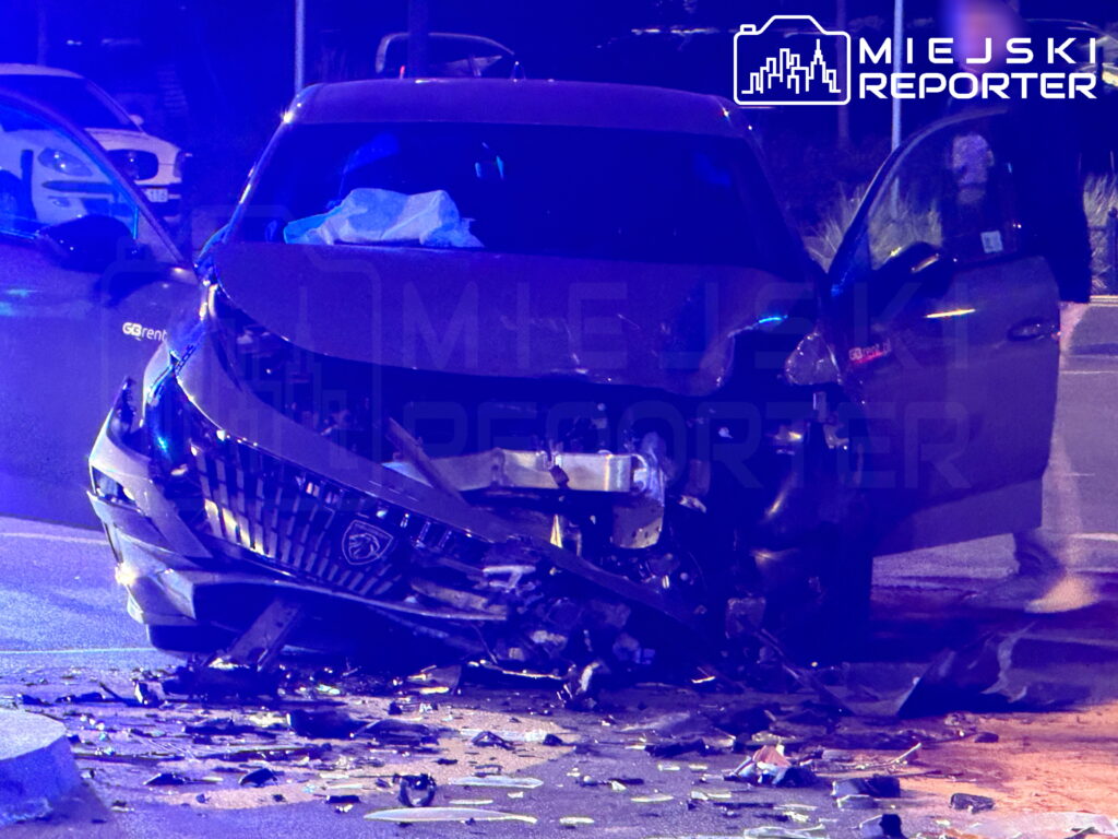Uszkodzony Peugeot z rozbitym przodem stoi nocą na ulicy, wokół leżą odłamki karoserii.
