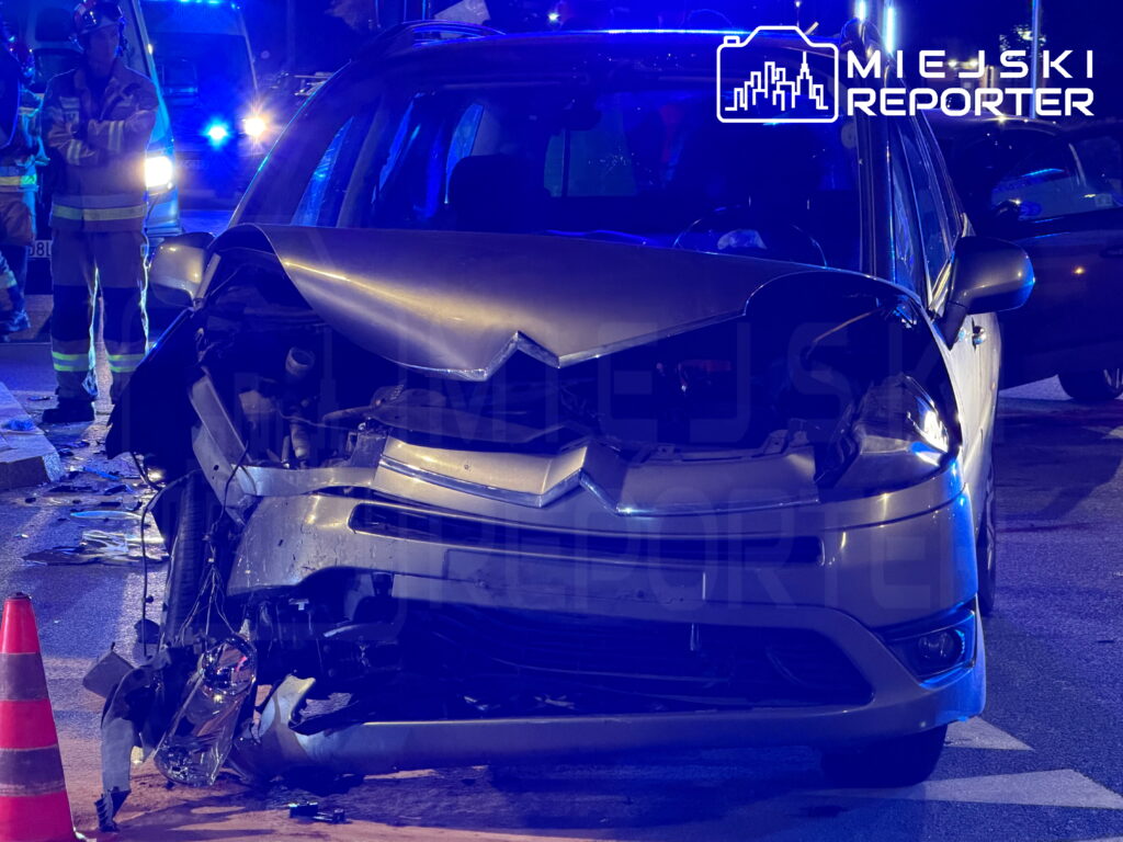 Uszkodzony samochód z rozbitą maską i przednim zderzakiem stoi na drodze nocą, obok pracuje ratownik w kamizelce odblaskowej.