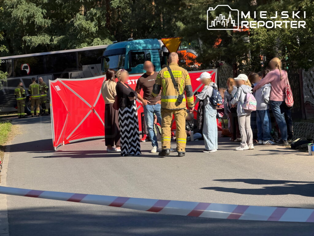 Strażak rozmawia z dorosłymi i dziećmi obok czerwonego parawanu, za którym stoją autokary; teren odgrodzono taśmą.