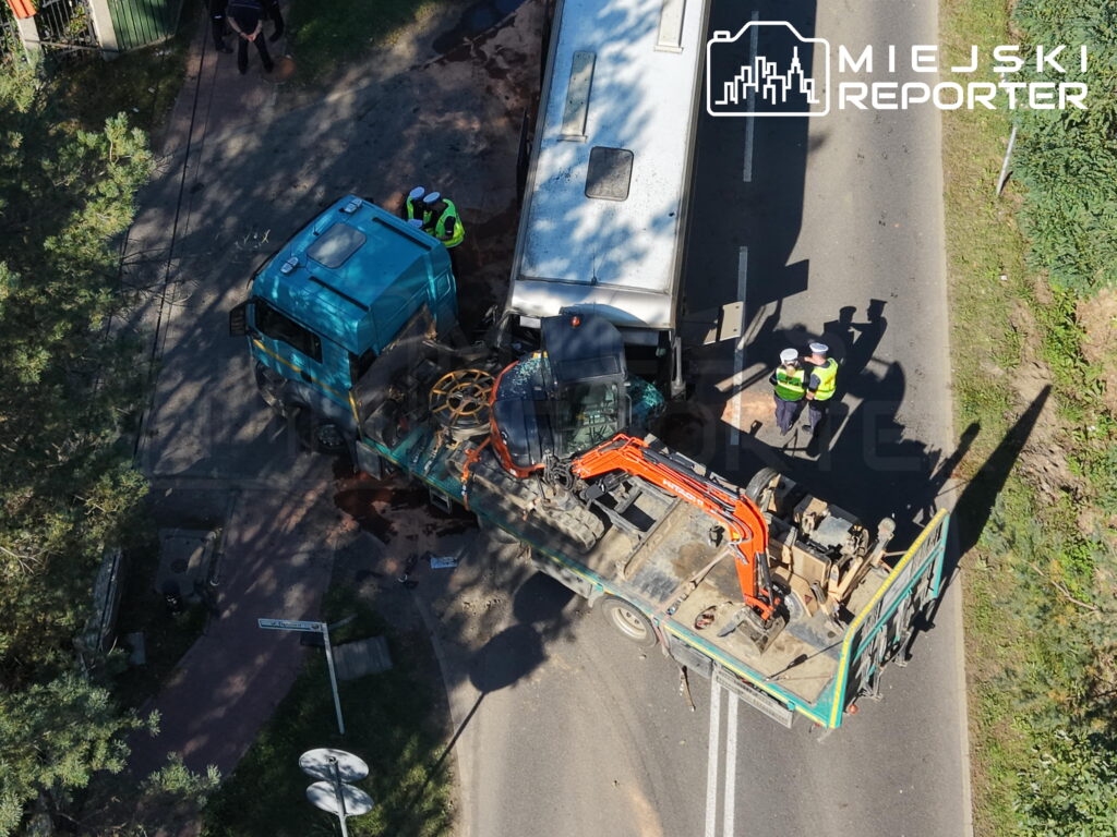 Ciężarówka z przyczepą przewożącą koparkę zderzyła się z autokarem na drodze; obok stoją służby ratunkowe.