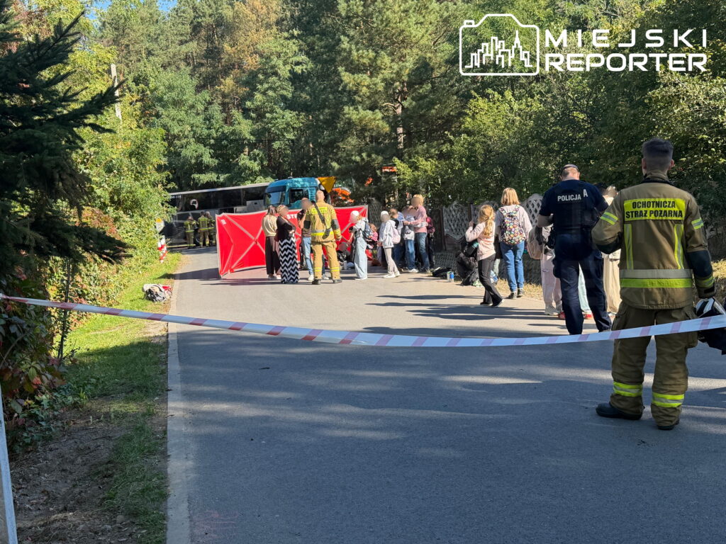 Policjant i strażak stoją za taśmą zabezpieczającą drogę, w tle służby ratunkowe i autokar pośród drzew.