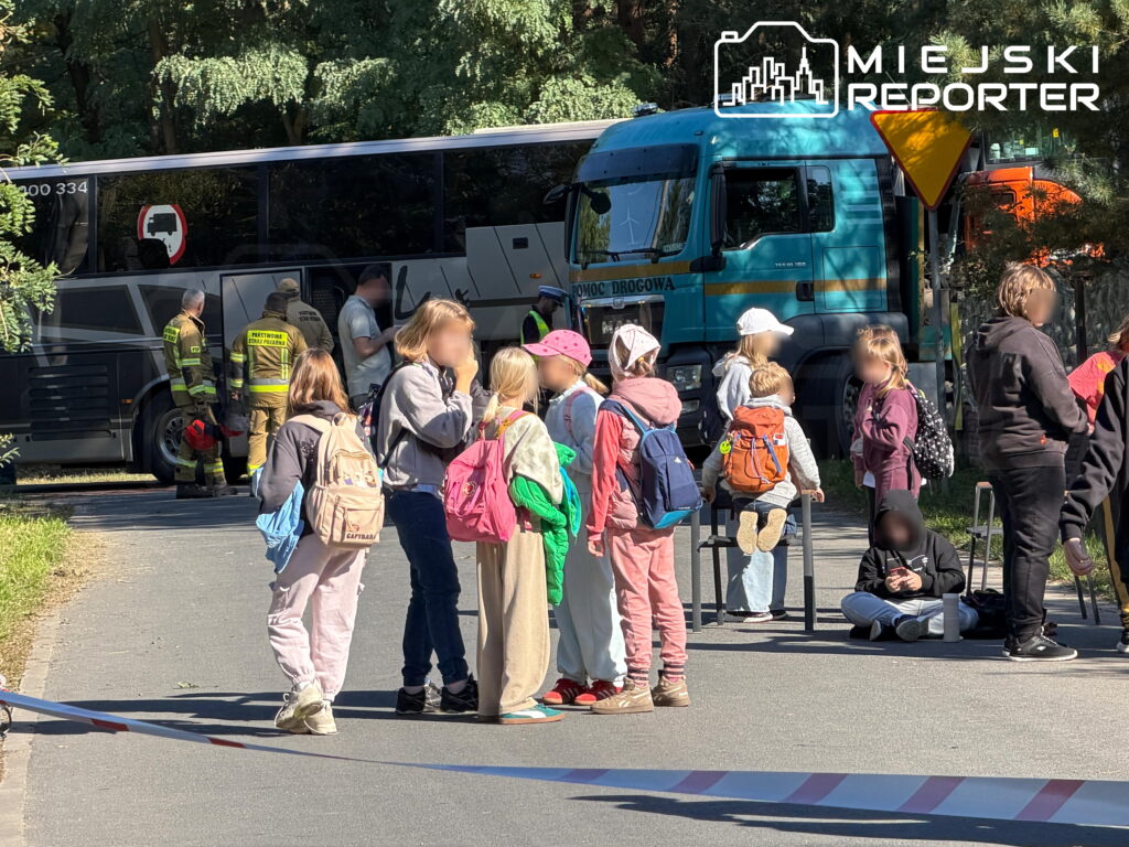 Dzieci z plecakami stoją przed autokarem i ciężarówką, obok których pracują służby ratunkowe.