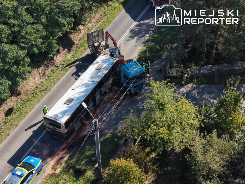 Autokar i niebieska ciężarówka po zderzeniu na wąskiej drodze, obok pracują służby ratunkowe.