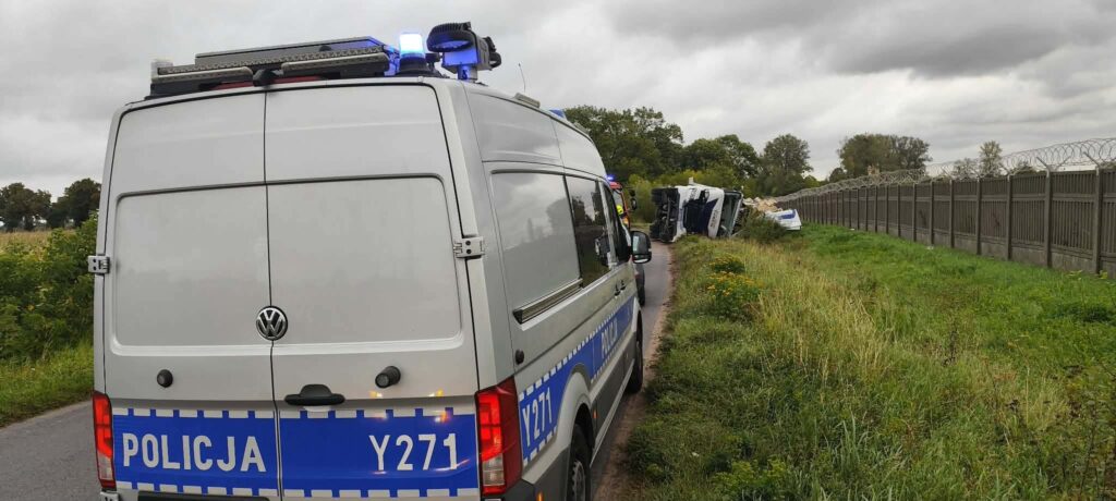 Policyjny radiowóz z niebieskimi światłami stoi obok przewróconego pojazdu na poboczu drogi, w tle ogrodzenie z drutu kolczastego.