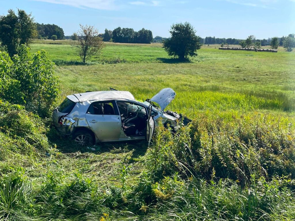 Zniszczony samochód osobowy znajduje się w wysokiej trawie na skraju pola, otoczony drzewami i krzewami.