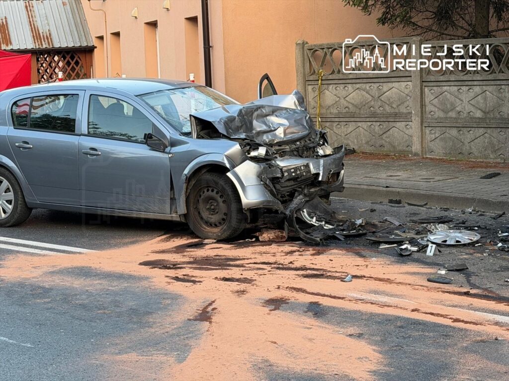 Zniszczony samochód osobowy leży na jezdni, obok rozrzucone fragmenty blachy i plamy płynów.