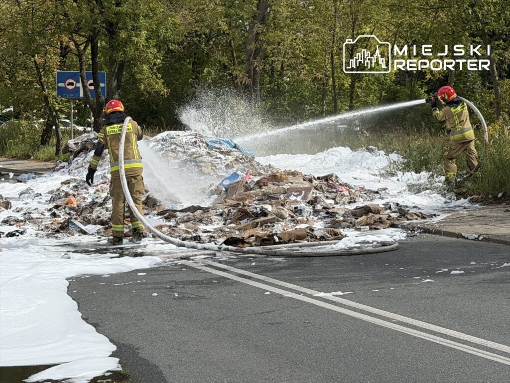 Strażacy w mundurach gaśniczych polewają wodą płonące odpady na jezdni, obok widoczna piana gaśnicza.