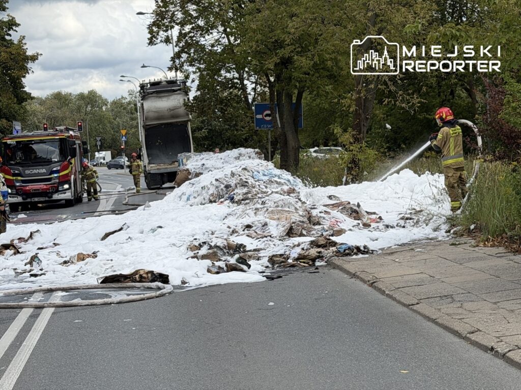 Strażacy gaszą pożar odpadów na jezdni, obok ciężarówki z uniesioną paką, pokrytej pianą gaśniczą.