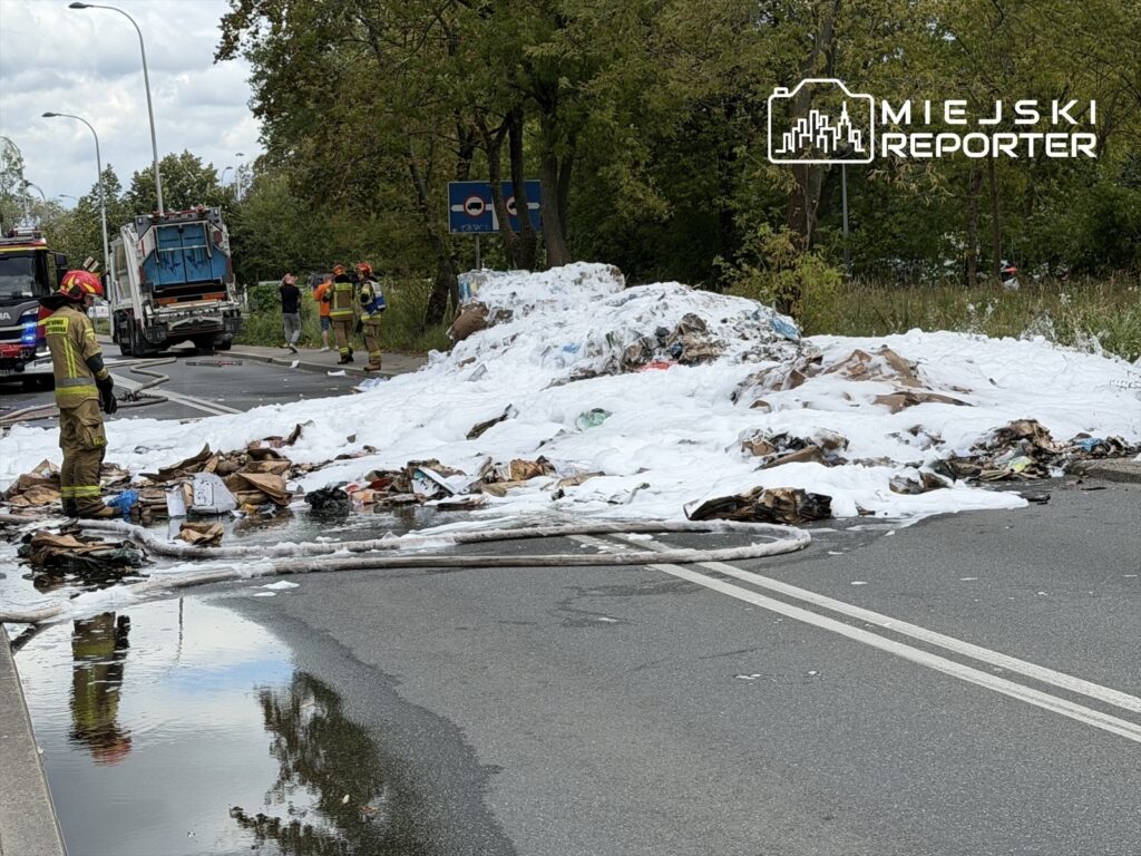 Strażacy gaszą płonące odpady na jezdni, obok leży pokryta pianą sterta śmieci, w tle widoczny wóz strażacki.