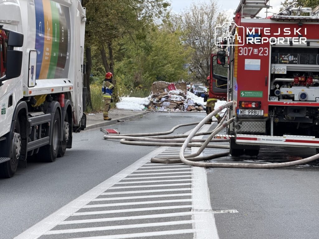 Strażak w mundurze obserwuje akcję gaśniczą przy płonących odpadach na jezdni, obok stoi wóz strażacki.