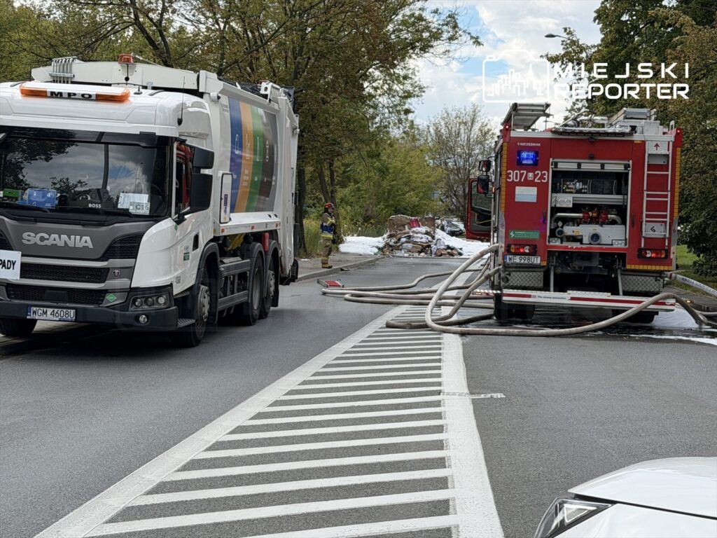 Strażacy gaszą pożar odpadów na jezdni, obok stoi ciężarówka z odpadami oraz wóz strażacki z rozwiniętymi wężami.