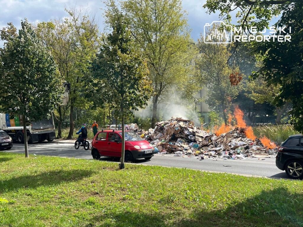 Płonące odpady wysypane na jezdnię, obok przejeżdżający rowerzysta i samochody w tle.