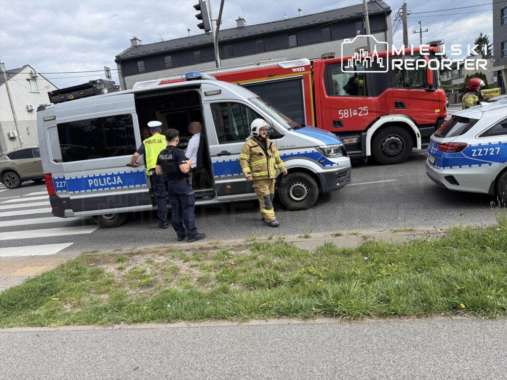 Policjant w mundurze prowadzi osobę do radiowozu, obok stoi wóz strażacki oraz drugi radiowóz na ruchliwej ulicy.