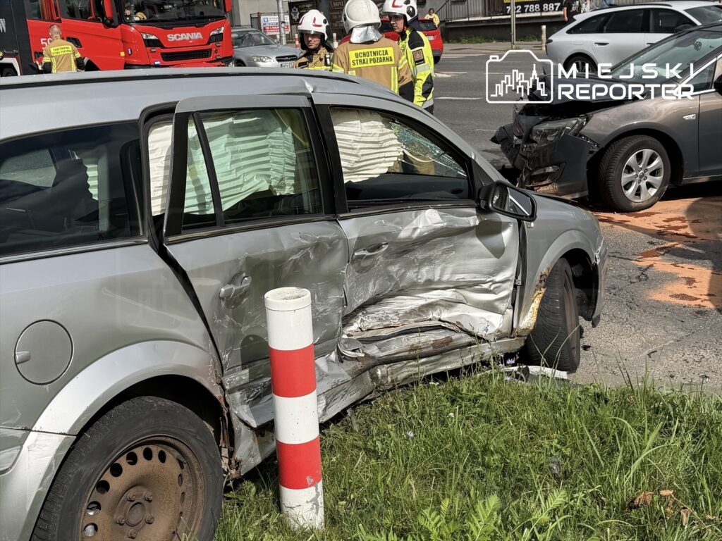 Uszkodzony samochód osobowy z wgniecioną stroną stoi obok pasów ruchu, a w tle widać służby ratunkowe i drugi pojazd po kolizji.