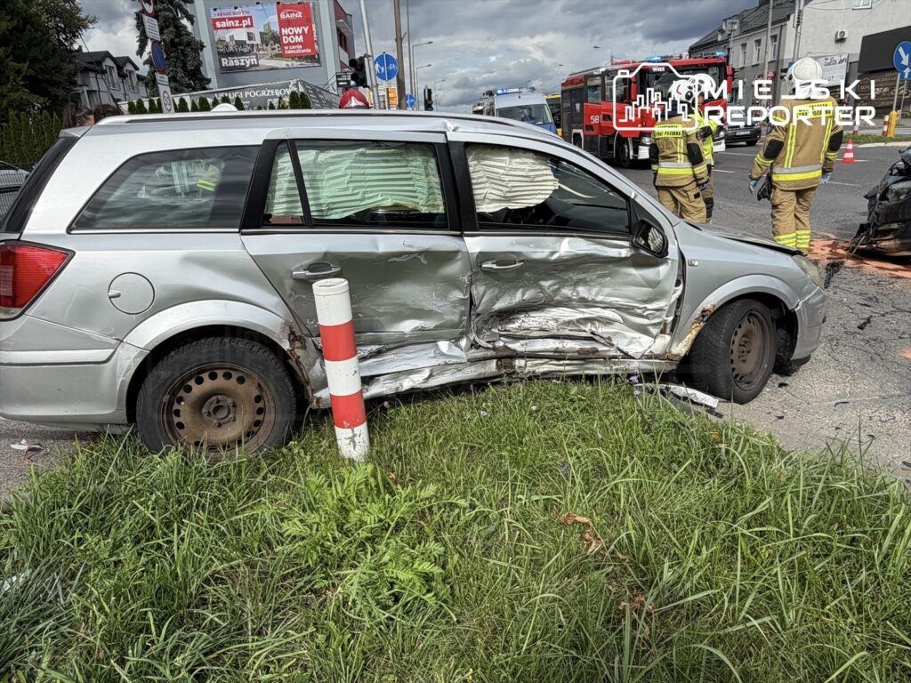 Uszkodzony samochód osobowy z wgniecioną boczną częścią stoi na poboczu, a obok interweniują strażacy.