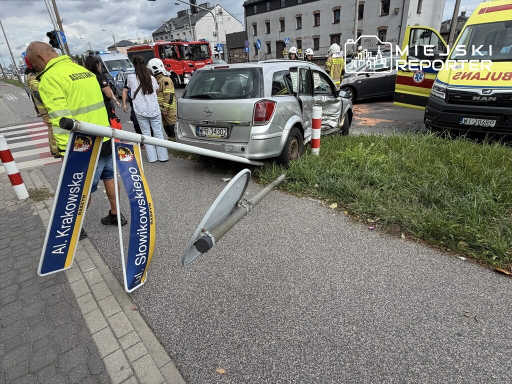 Kierowca z uszkodzonym samochodem stoi obok przewróconego znaku drogowym na ulicy, otoczony przez służby ratunkowe.