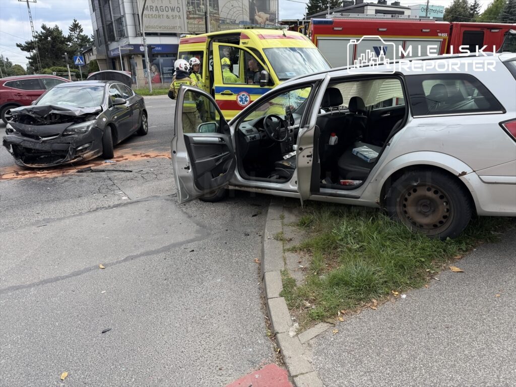 Czarny samochód osobowy z uszkodzoną przednią częścią stoi na jezdni, obok otwarty pojazd z widocznymi śladami kolizji.