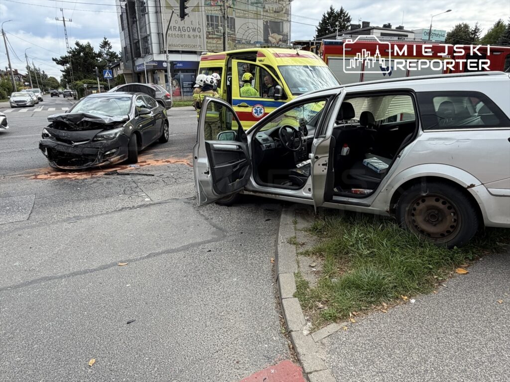 Czarne auto z uszkodzonym przodem stoi na skrzyżowaniu, obok białe auto z otwartymi drzwiami, w tle karetka.