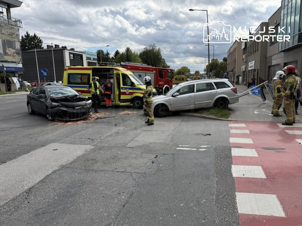 Czarny samochód osobowy z uszkodzoną przednią częścią stoi na jezdni, obok niego zespół ratunkowy oraz strażacy zabezpieczają miejsce kolizji. Na poboczu leży zniszczony znak drogowy, a w oddali widoczny jest czerwony wóz strażacki.