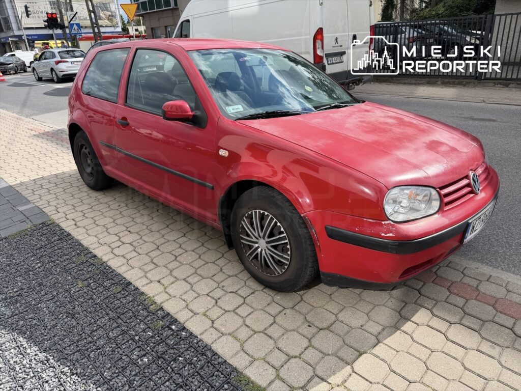 Czerwony samochód osobowy marki Volkswagen zaparkowany na chodniku, obok ruchliwej ulicy z widocznymi innymi pojazdami.