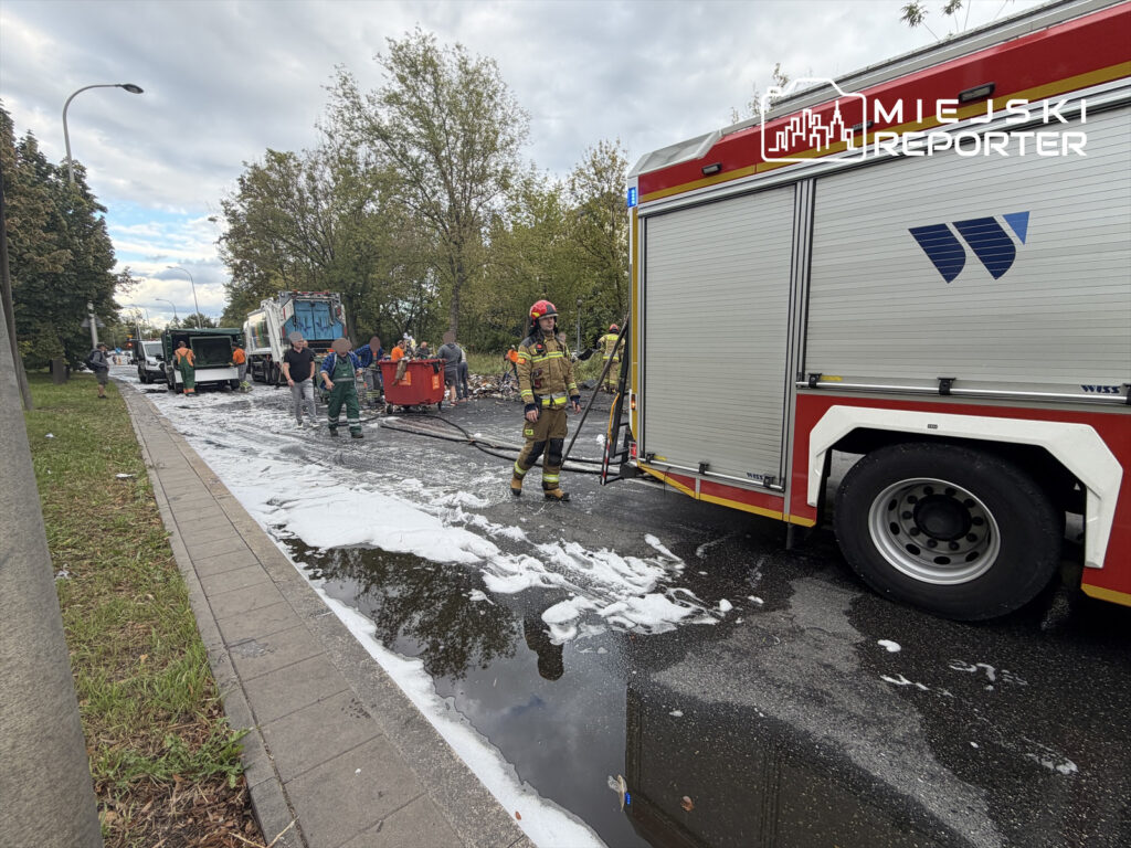 Grupa strażaków i pracowników sprzątających usuwa pianę z jezdni po kolizji, obok stoi wóz strażacki.