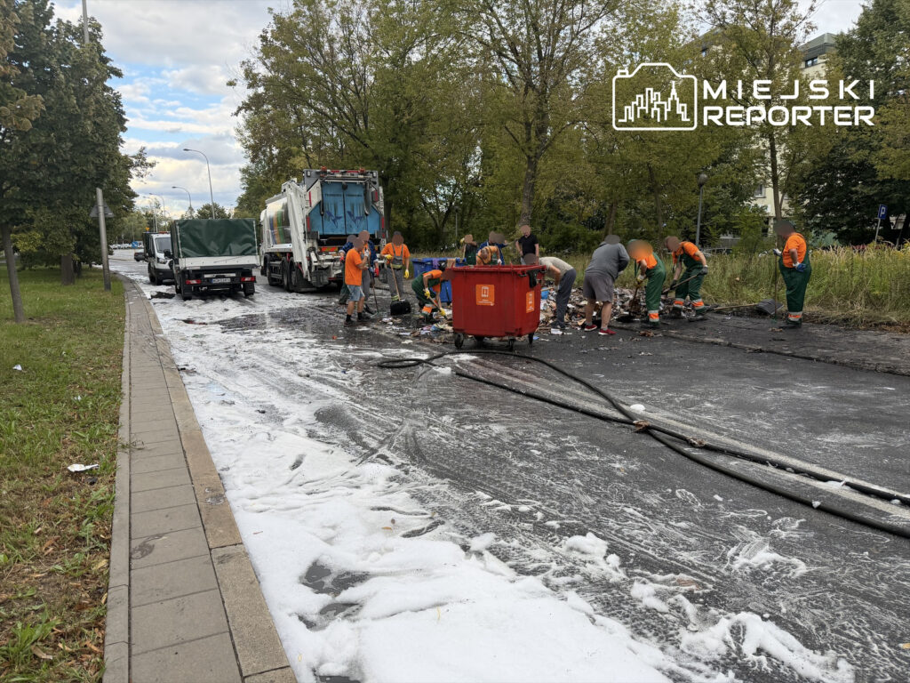 Grupa pracowników w pomarańczowych koszulach sprząta rozlaną ciecz na drodze, obok stoją pojazdy komunalne.