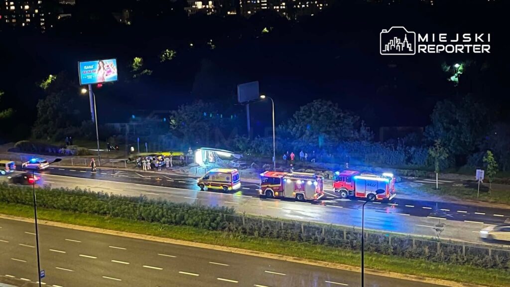 Na drodze w nocy, pojazdy służb ratunkowych interweniują przy przewróconym autobusie, a w tle widoczne są policyjne sygnały świetlne.