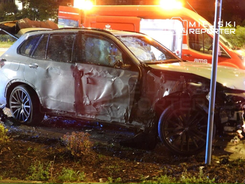 Uszkodzone BMW stoi na chodniku, obok widać woz ambulansowy z włączonymi sygnałami świetlnymi.