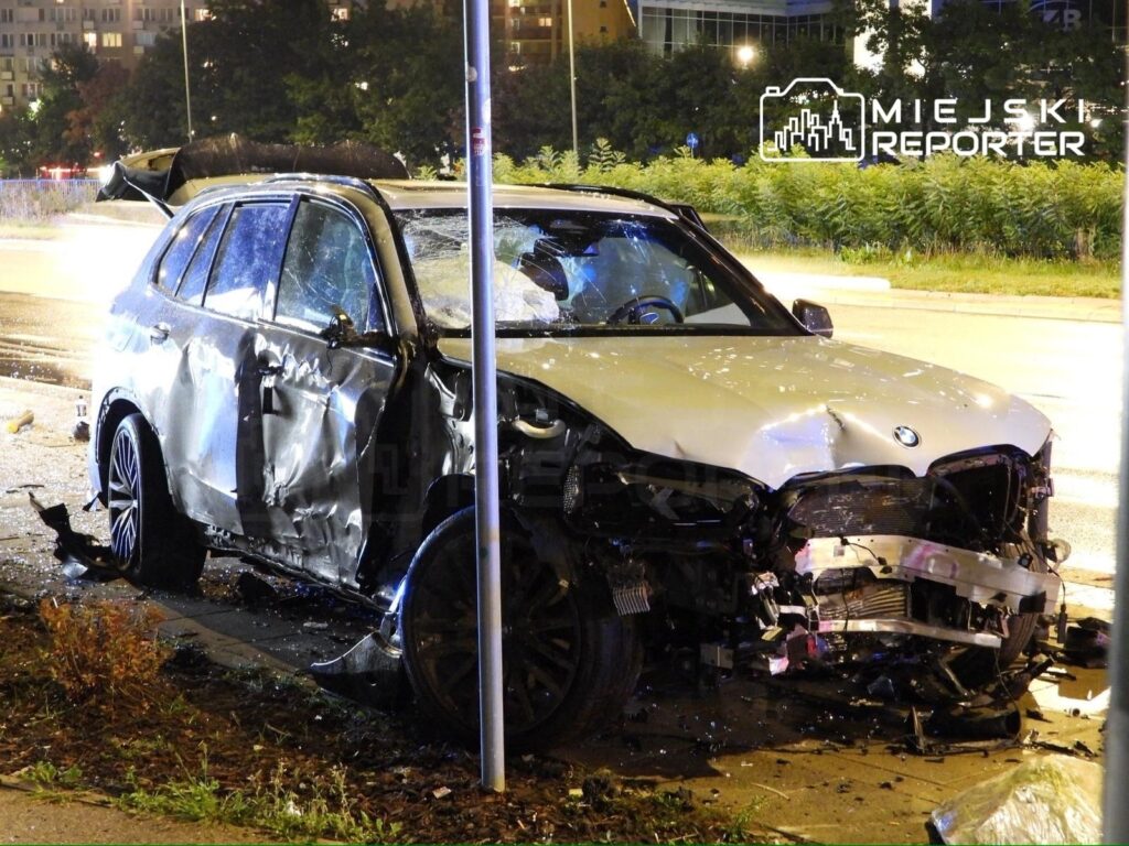 Zniszczony samochód marki BMW stoi na chodniku po wypadku, z widocznymi uszkodzeniami nadwozia i zniszczonym przodem.