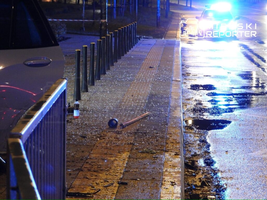 Na chodniku leżą kawałki szkła, obok widać przewróconą niebieską piłkę i fragmenty drewnianego słupa, w tle radiowóz z włączonymi sygnałami świetlnymi.