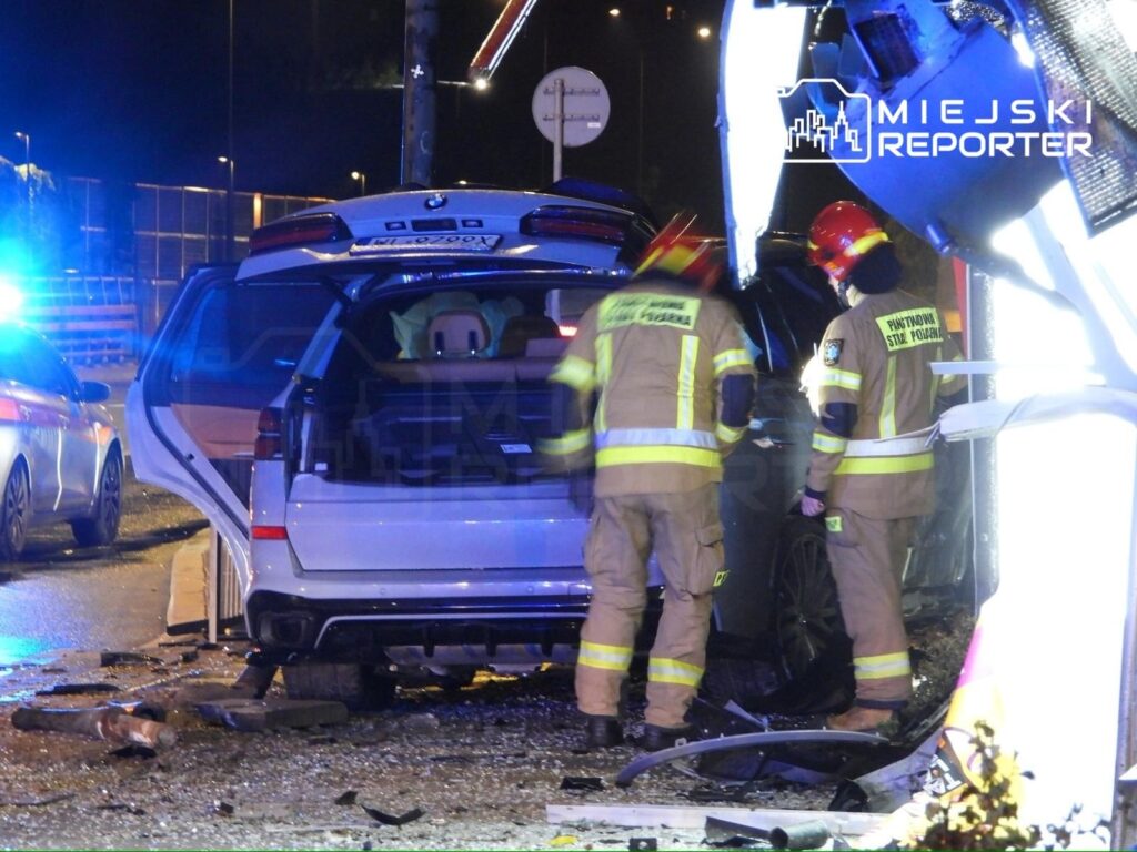 Biały samochód BMW uszkodzony po wypadku, obok dwóch strażaków w mundurach, udzielających pomocy.