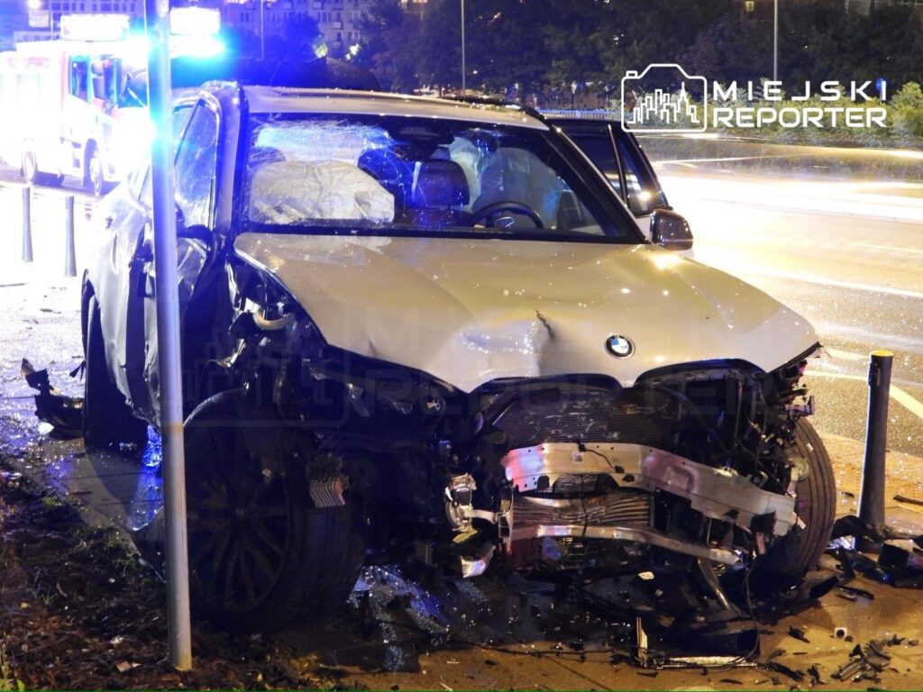 Zniszczone BMW z uszkodzoną przednią częścią stoi przy przystanku, obok widać niebieskie światła służb ratunkowych.